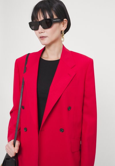 Blazer rouge croisé avec des boutons noirs, porté sur un haut noir. Les accessoires comprennent de grandes lunettes de soleil noires et des boucles d'oreilles en chaîne dorée.
