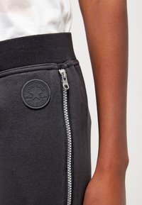 Schwarze Jogginghosen mit einem gerippten Bund, ausgestattet mit einem seitlichen Reißverschluss und einem runden schwarzen Gummiemblem mit dem Converse-Logo.
