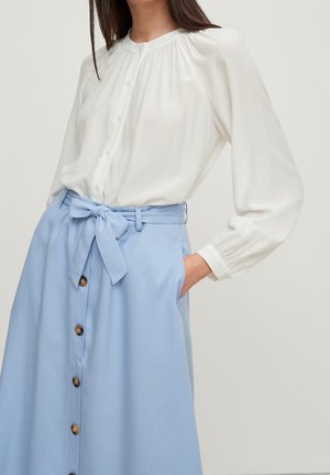 Femme portant un chemisier blanc à manches longues boutonné, rentré dans une jupe bleu clair taille haute avec des boutons devant et une ceinture nouée.