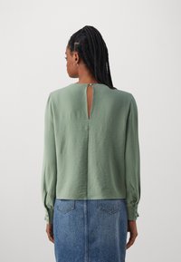 Blusa verde de manga larga con cuello redondo y detalle de ojal en la parte posterior. Presenta una tela suave y texturizada con puños abotonados. Combinada con una falda de mezclilla.