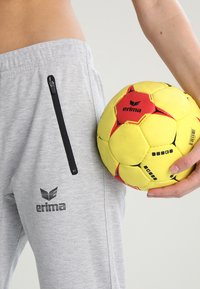 Pantalones de chándal grises con un bolsillo con cremallera negra, que presenta el logo "erima". Se sostiene una pelota de balonmano amarilla brillante con acentos rojos y negros.