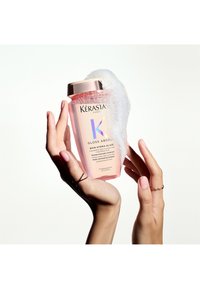 Kérastase Gloss Absolu shampoofles, roze met een gouden dop, vastgehouden in handen, omringd door bellen, met een prominent logo en productdetails.