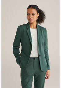 Žalias pritaikytas švarkas, derinamas su atitinkančiomis kelnėmis. Švarkas turi įpjautą apykaklę, lygų audinį ir minimalizmo stiliaus detales.