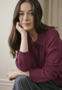 Pulover tricotat burgund cu guler și trei butoane negre. Textură moale. Păr mătăsos ce cade pe umeri, cu un fundal neutru.