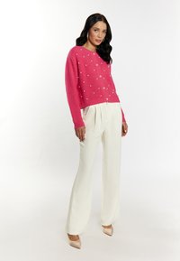 Maglione rosa lavorato a maglia con piccole perle decorative, abbinato a pantaloni larghi color avorio e tacchi nudi. La modella posa su uno sfondo bianco.