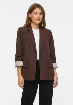 Mulher a usar um blazer castanho com punhos às riscas dobrados sobre uma t-shirt branca e calças pretas, em pé contra um fundo claro e simples.