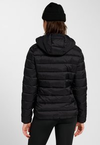 Czarna kurtka puchowa z kapturem, z pikowanymi panelami, gładką fakturą i elastycznymi mankietami. Osoba nosząca kurtkę ubrana jest w czarne legginsy.
