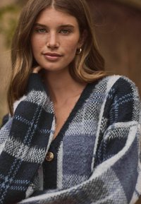 Cardigan en laine avec un motif à carreaux bleu et gris, décolleté en V, et fermeture à boutons visible, avec des manches en tissu texturé et doux.
