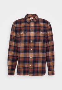Chemise en flanelle avec un motif à carreaux multicolore dans des teintes de marron, bleu et beige. Elle a un col boutonné et deux poches poitrine à boutons.