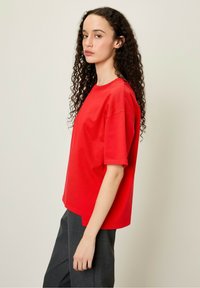 Raudona medvilninė marškinėlė su trumpomis rankovėmis, apvaliu iškirpimu ir laisvu kirpimu, derinama su pilkomis kelnėmis. Matomi garbanoti plaukai.