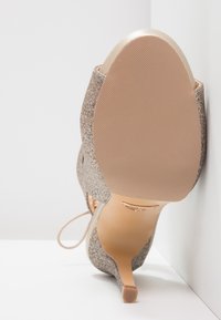 Sepatu hak tinggi berwarna beige yang berkilau dengan ujung runcing, aksen emas halus pada tumit, dan sol bertekstur untuk pegangan.