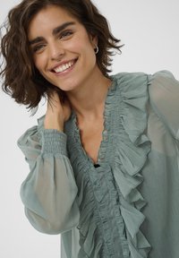Lichtgroene transparante blouse met rimpeldetails aan de voorkant, smock-mouwen en een losse pasvorm. De stof heeft een zachte textuur en een vloeiende uitstraling.