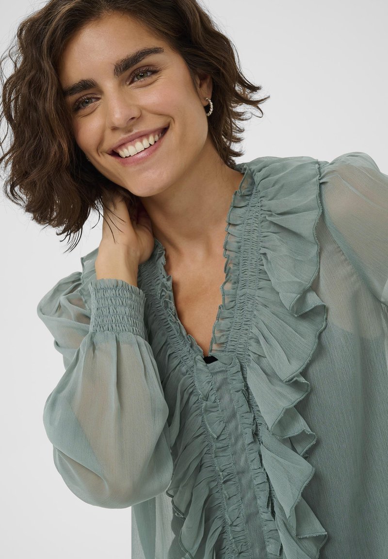 Blouse légère vert clair en tissu transparent avec des détails froncés à l'avant, des poignets smockés et une coupe ample. Le tissu a une texture douce et un design fluide.