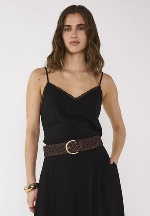 Femme aux cheveux bruns ondulés, portant un caraco noir à bordure de dentelle, un pantalon noir, une ceinture tressée marron et un bracelet doré, debout la main dans la poche.