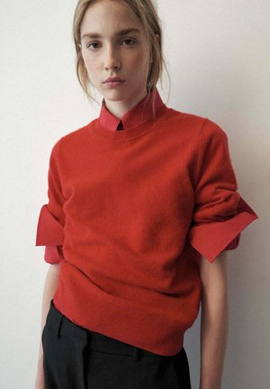 Joven mujer con cabello rubio lleva un suéter rojo sobre una camisa roja con cuello y mangas remangadas y pantalones negros, de pie contra una pared lisa y clara.