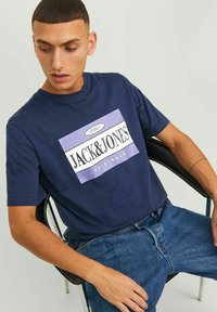 Camiseta de manga corta azul marino con un gran gráfico en blanco y lavanda que incluye el texto "JACK & JONES ORIGINALS", con cuello redondo.
