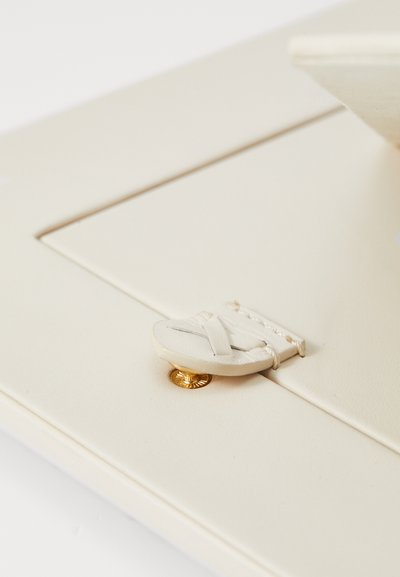 Accessorio in pelle bianca con chiusura a bottone rotondo caratterizzato da un design intrecciato. Elementi in metallo color oro accentuano il materiale in pelle.