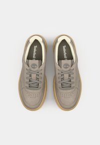 Adidași din suede gri deschis cu o talpă din cauciuc bej, dotati cu șireturi gri, detalii perforate la vârf și o etichetă cu logo pe limbă.