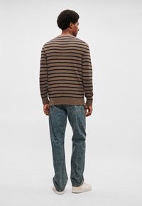 Καφέ και ναυτικό ριγέ πλεκτό πουλόβερ με στρογγυλή λαιμόκοψη, συνδυασμένο με φαρδιά, ξεθωριασμένα μπλε τζιν και ανοιχτόχρωμα αθλητικά παπούτσια.