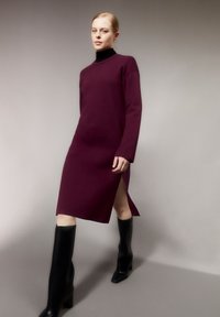 Abito in maglia borgogna con maniche lunghe e uno spacco alto, abbinato a stivali neri alti fino al ginocchio con tacco a spillo, che mettono in risalto una texture liscia.
