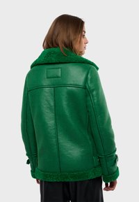 Zelená kožená bunda s huňatým zeleným koženým golierom, textúrovaný povrch a nastaviteľné pásy na rukávoch. Jednoduchý, štruktúrovaný dizajn.