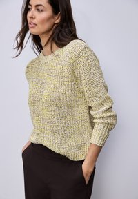 Femme portant un pull en maille texturé jaune et beige à manches longues et un pantalon noir, les mains dans les poches, regardant légèrement vers la gauche.