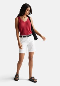 Top senza maniche rosso, pantaloni corti bianchi con risvolto, cintura nera e sandali neri con dettagli. Il modello tiene una borsa a spalla nera strutturata.