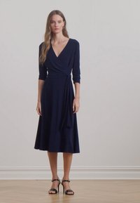 Abito a portafoglio blu navy con maniche a tre quarti, gonna fluida, tessuto morbido e dettaglio con laccetto in vita, abbinato a sandali neri con tacco alto.