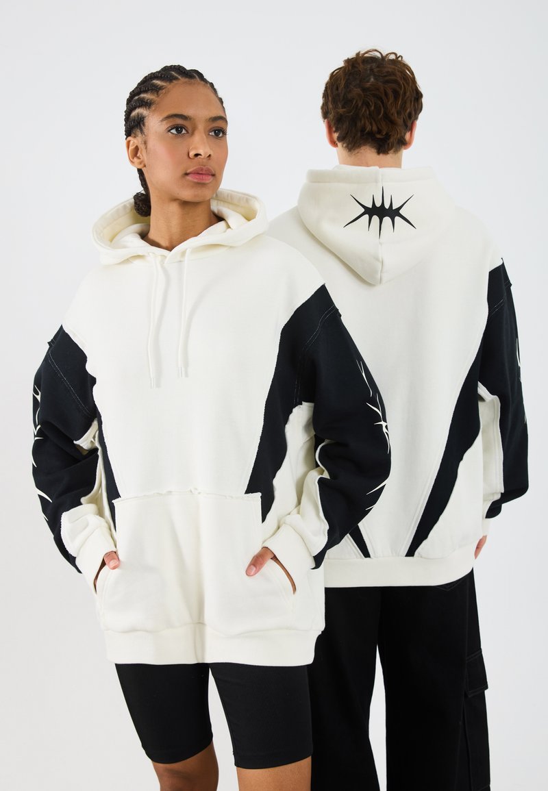 Witte en zwarte hoodie met een capuchon, voorkruiden en een grafisch ontwerp op de zwarte mouwen en achterkant. Gemaakt van zachte, gestructureerde stof.