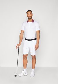 Bijela golf polo majica s uzorkastim crvenim ovratnikom, uparena s bijelim kratkim hlačama. Model drži golf palicu, odjeven u bijele tenisice i čarape.
