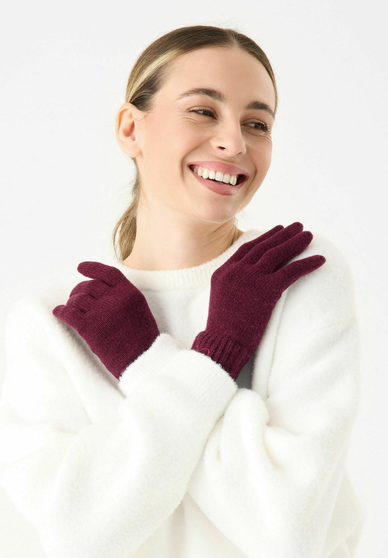Gants en tricot bordeaux avec des poignets côtelés, présentés sur un pull blanc. Les gants sont d'une texture douce et s'adaptent parfaitement.