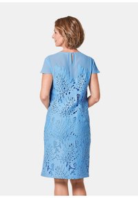 Helles blaues Kleid mit kurzen Ärmeln, einem durchsichtigen oberen Abschnitt und kunstvollen blattförmigen Ausschnitten auf dem Rücken und dem Rock.
