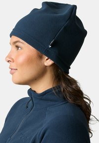 Beanie in pile blu navy con design scivolato, caratterizzato da un accento laterale e tessuto texturizzato. Indossato con un top in pile a collo alto abbinato.