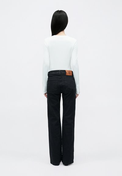 Jeans noirs à jambes larges avec un patch logo en cuir marron à l'arrière, associés à un haut blanc ajusté à manches longues.