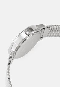 Montre en maille argentée avec un boîtier rond, une couronne texturée et un dos lisse. Le bracelet présente un design tissé, améliorant la flexibilité et le confort.