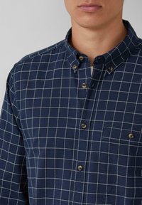Camicia a quadri blu navy in cotone. Caratterizzata da un colletto con bottoni, taschino sul petto e bottoni marroni con motivo a griglia bianca.