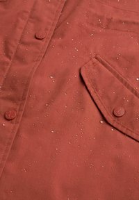 Vattentålig rost-röd jacktyg med synliga vattendroppar, utrustad med knäppning och bröstficka. Slät textur.