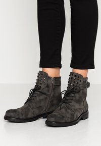 Bottines en daim gris avec un bout arrondi, un laçage à l'avant, une fermeture éclair latérale et des accents cloutés sur le col. Semelle en caoutchouc noire.