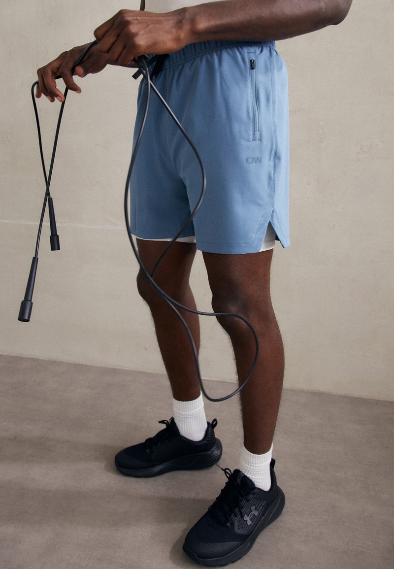 Shorts de sport bleu clair avec des fentes sur les côtés, associés à des baskets noires et des chaussettes blanches montantes. Une corde à sauter est tenue dans les mains.