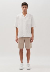 Witte korte mouwen overhemd met een enkele borstzak, gecombineerd met beige opgerolde shorts en witte sneakers op een neutrale achtergrond.
