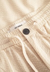 Heledad beežid püksid tekstuurse kangaga, elastse vöökohaga ja musta nööbiga kinnitusega. Sildil on kiri "GABBA" koos suuruseinfoga.