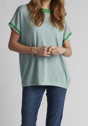 Vrouw draagt een lichtblauwe losgebreide top met groene rand en donkerblauwe spijkerbroek, handen voor elkaar geklemd, effen grijze achtergrond.