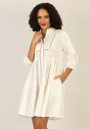 Woman with curly hair wearing a white, knee-length dress with three-quarter sleeves and decorative pintuck and cutout details.