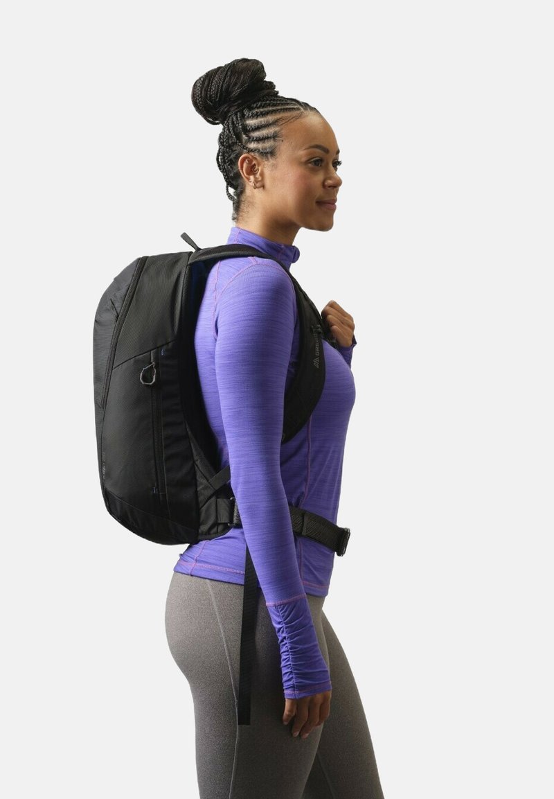 Femme aux cheveux tressés portant un sac à dos noir, un haut à manches longues violet et des leggings gris, debout de profil sur un fond blanc.