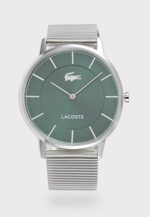 Montre à cadran vert avec boîtier en argent et bracelet en métal strié. Arbore un logo Lacoste, des index simples et un design épuré. Forme ronde, style minimaliste.