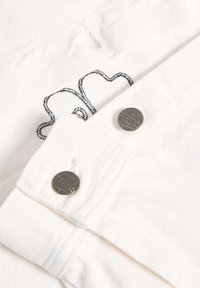 Camisa de tela blanca con diseño bordado en negro. Dos botones redondos plateados en los puños. Texturizada con un sutil patrón de tejido.