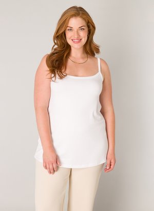 Lächelnde Frau mit langen roten Haaren, die ein weißes ärmelloses Oberteil und beige Hosen trägt, vor einem schlichten hellen Hintergrund stehend.