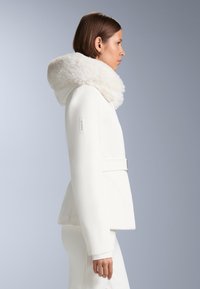 Jachetă albă cu glugă căptușită cu blană, croială ajustată, suprafață texturată, talie cu curea și branding subtil pe mânecă.