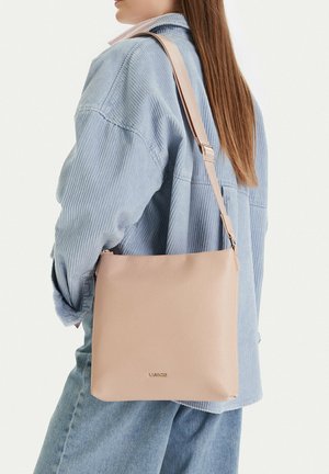 Mujer con chaqueta de pana azul claro que lleva un bolso de cuero beige para el hombro con correa ajustable y logo sutil.