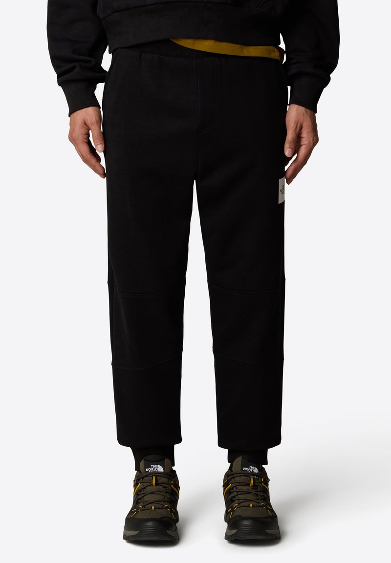 Czarne dresy o luźnym kroju, z elastycznym pasem, kieszenią naszywaną na biodrze oraz teksturowanym materiałem, w połączeniu z butami trekkingowymi.